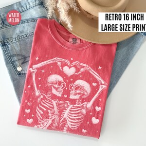 May include: A red t-shirt with a graphic of two skeletons wearing heart-shaped sunglasses, forming a heart with their arms. The shirt is labeled "Retro 16 inch Large Size Print". The shirt is placed on top of blue jeans and a tan hat.