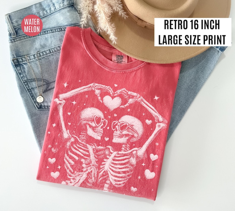 May include: A red t-shirt with a graphic of two skeletons wearing heart-shaped sunglasses, forming a heart with their arms. The shirt is labeled "Retro 16 inch Large Size Print". The shirt is placed on top of blue jeans and a tan hat.