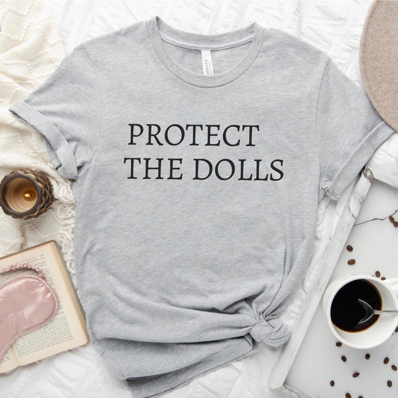 Puede incluir: Una camiseta gris con el texto "PROTECT THE DOLLS" impreso en negro.