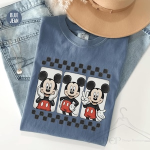 Puede incluir: Una camiseta azul con tres im&aacute;genes de Mickey Mouse en diferentes poses, cada una enmarcada por un patr&oacute;n de cuadros blancos y negros. La camiseta se muestra con vaqueros azules y un sombrero beige.