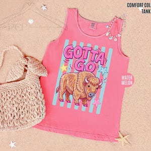 K&ouml;nnte beinhalten: Ein pinkfarbenes Tanktop mit den Worten "GOTTA GO" in einer Blasen-Schriftart, einem Cartoon-Bison und Sternen. Das Tanktop befindet sich neben einer gewebten beigefarbenen Handtasche.