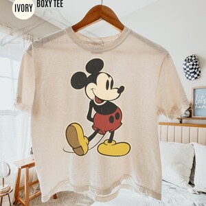 May include: Ivory boxy tee with a vintage Mickey Mouse graphic. The cartoon character is in black, red, yellow, and beige. The shirt hangs on a wooden hanger. Text on the shirt reads "COMFORT COLORS IVORY BOXY TEE."