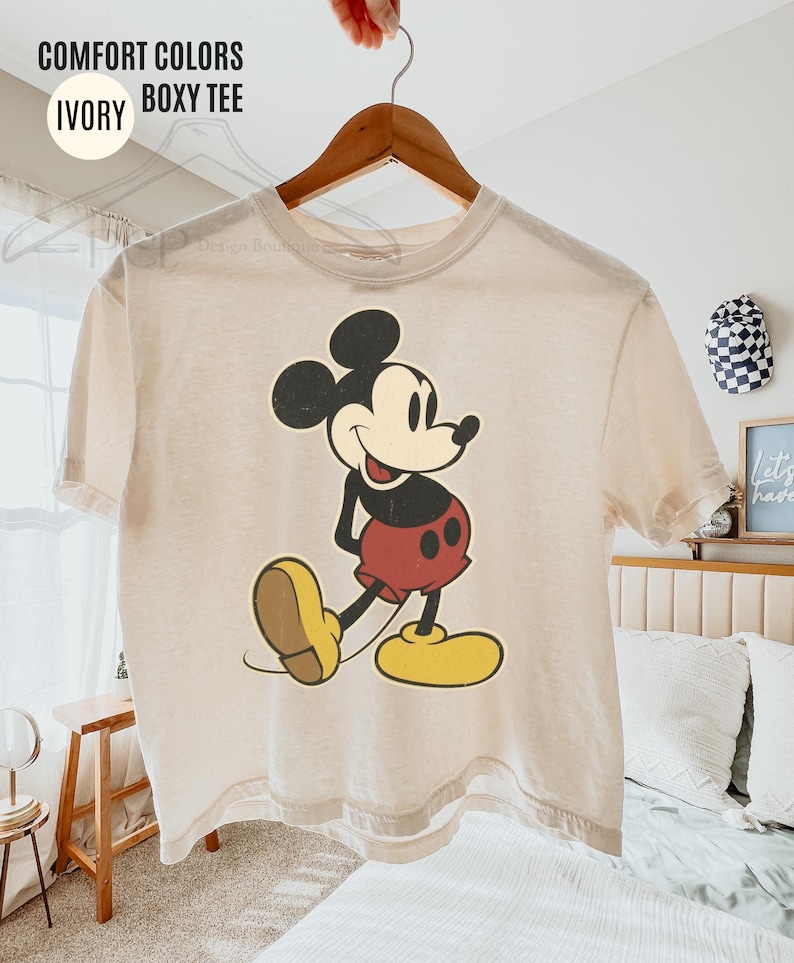 May include: Ivory boxy tee with a vintage Mickey Mouse graphic. The cartoon character is in black, red, yellow, and beige. The shirt hangs on a wooden hanger. Text on the shirt reads "COMFORT COLORS IVORY BOXY TEE."