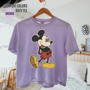 May include: Orchid-coloured boxy tee featuring a vintage-style Mickey Mouse graphic. The cartoon character is depicted in black, red, yellow, and tan. The shirt has the words "COMFORT COLORS" and "ORCHID BOXY TEE" printed on it.