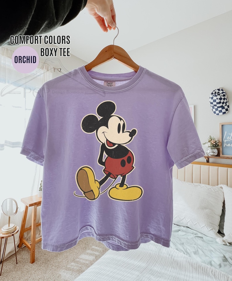 May include: Orchid-coloured boxy tee featuring a vintage-style Mickey Mouse graphic. The cartoon character is depicted in black, red, yellow, and tan. The shirt has the words "COMFORT COLORS" and "ORCHID BOXY TEE" printed on it.