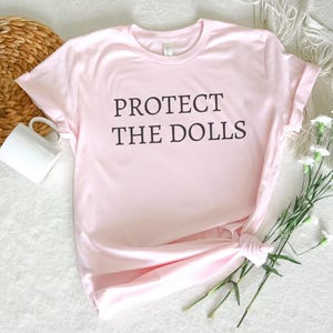 Puede incluir: Una camiseta rosa con el texto "PROTECT THE DOLLS" impreso en negro.