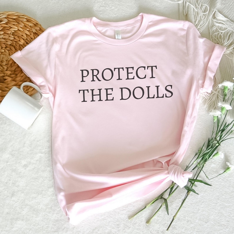 Puede incluir: Una camiseta rosa con el texto "PROTECT THE DOLLS" impreso en negro.
