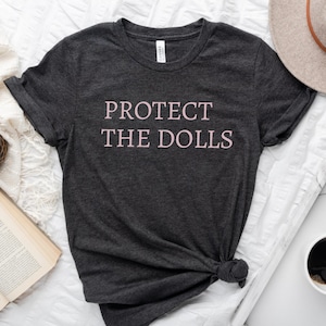 Puede incluir: Una camiseta gris oscuro con el texto "PROTECT THE DOLLS" impreso en rosa.