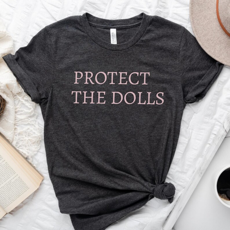 Puede incluir: Una camiseta gris oscuro con el texto "PROTECT THE DOLLS" impreso en rosa.