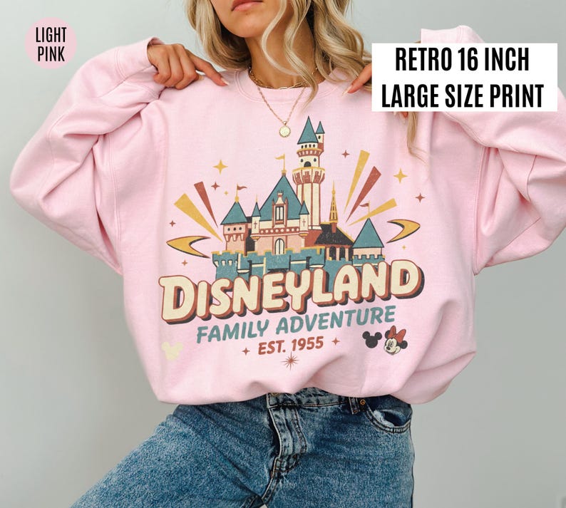 May include: Light pink sweatshirt with a retro Disneyland graphic. The design features a castle, the word "Disneyland", and the text "Family Adventure Est. 1955". The print is 16 inches in size.