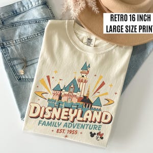 Puede incluir: Camiseta color crema con un dise&ntilde;o gr&aacute;fico retro de Disneyland. El dise&ntilde;o presenta un castillo, la palabra "Disneyland" y "Family Adventure + EST. 1955 +" en un estilo vintage. La camiseta se combina con unos vaqueros azules y un sombrero beige.