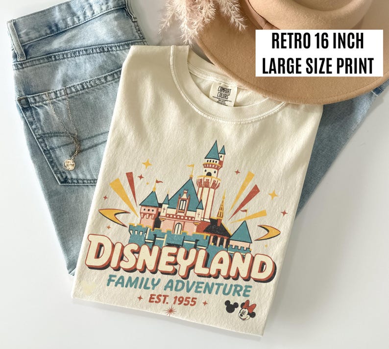 Puede incluir: Camiseta color crema con un dise&ntilde;o gr&aacute;fico retro de Disneyland. El dise&ntilde;o presenta un castillo, la palabra "Disneyland" y "Family Adventure + EST. 1955 +" en un estilo vintage. La camiseta se combina con unos vaqueros azules y un sombrero beige.