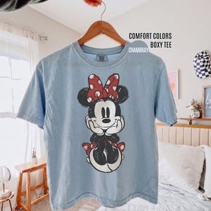 May include: A light blue Comfort Colors boxy tee shirt featuring a graphic of Minnie Mouse. Minnie is depicted in black and white with a red bow and accents. The shirt is hanging on a wooden hanger.