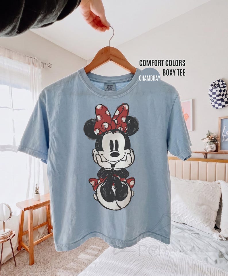 May include: A light blue Comfort Colors boxy tee shirt featuring a graphic of Minnie Mouse. Minnie is depicted in black and white with a red bow and accents. The shirt is hanging on a wooden hanger.