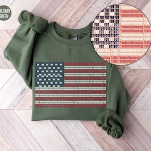May include: Military green sweatshirt featuring an American flag design. The flag is composed of text blocks with the words "Freedom Democracy Truth" and "FDT". The sweatshirt has a crew neck and long sleeves.