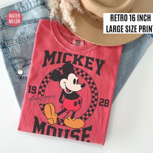 May include: A red t-shirt featuring a graphic of Mickey Mouse. The shirt has the words "Mickey Mouse" in large black letters. The shirt is paired with blue jeans and a tan hat. The text "Retro 16 inch Large Size Print" is visible.