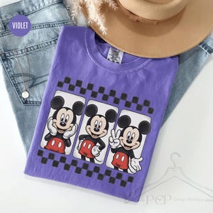 Puede incluir: Una camiseta violeta con tres im&aacute;genes de Mickey Mouse en rect&aacute;ngulos blancos con un borde a cuadros en blanco y negro. Mickey lleva pantalones cortos rojos y guantes blancos. La camiseta est&aacute; sobre unos vaqueros azules y un sombrero marr&oacute;n.