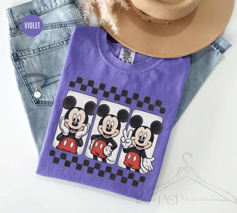 Puede incluir: Una camiseta violeta con tres im&aacute;genes de Mickey Mouse en rect&aacute;ngulos blancos con un borde a cuadros en blanco y negro. Mickey lleva pantalones cortos rojos y guantes blancos. La camiseta est&aacute; sobre unos vaqueros azules y un sombrero marr&oacute;n.
