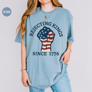 Puede incluir: Camiseta azul claro con el texto "Rejecting Kings Since 1776" y un dise&ntilde;o de pu&ntilde;o en los colores de la bandera estadounidense. La camiseta tiene cuello redondo y mangas cortas.