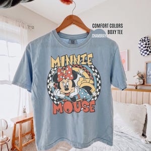 Może przedstawiać: Jasnoniebieska koszulka Comfort Colors z grafiką Myszki Minnie. Projekt przedstawia twarz Minnie z czerwoną kokardą w grochy, napisem "MINNIE MOUSE" i szachownicowym tłem. Koszulka wisi na drewnianym wieszaku.
