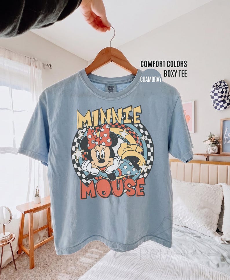 Może przedstawiać: Jasnoniebieska koszulka Comfort Colors z grafiką Myszki Minnie. Projekt przedstawia twarz Minnie z czerwoną kokardą w grochy, napisem "MINNIE MOUSE" i szachownicowym tłem. Koszulka wisi na drewnianym wieszaku.
