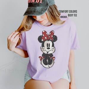 May include: A light purple Comfort Colors boxy tee featuring a Minnie Mouse graphic. The graphic shows Minnie Mouse with a red polka dot bow and red polka dot accents. The text "COMFORT COLORS BOXY TEE ORCHID" is also visible.
