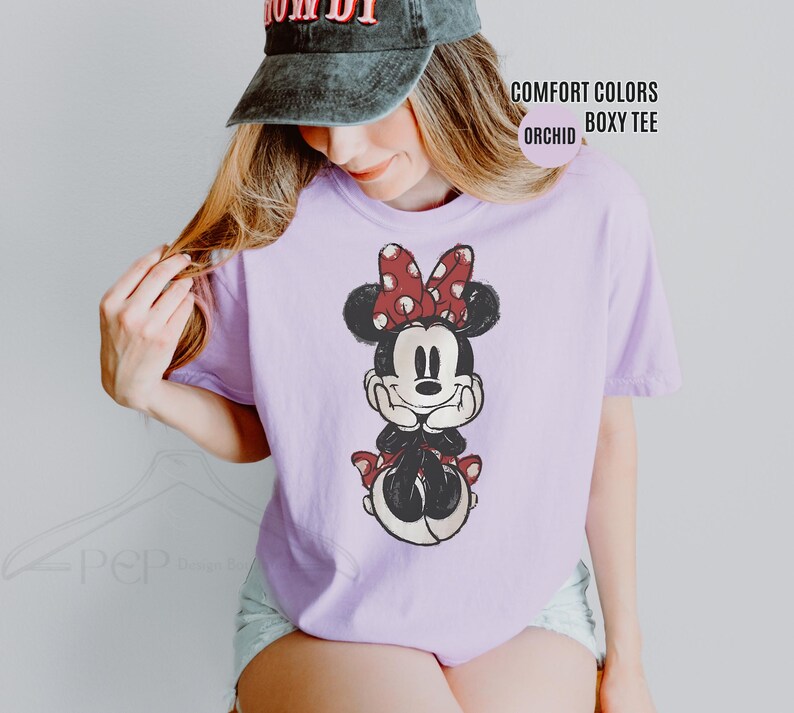 May include: A light purple Comfort Colors boxy tee featuring a Minnie Mouse graphic. The graphic shows Minnie Mouse with a red polka dot bow and red polka dot accents. The text "COMFORT COLORS BOXY TEE ORCHID" is also visible.