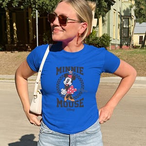 K&ouml;nnte beinhalten: Ein k&ouml;nigsblaues T-Shirt mit einem Minnie Mouse-Motiv. Das Design enth&auml;lt den Text "MINNIE MOUSE" in Schwarz, mit Minnie in einem gepunkteten Kleid. Das T-Shirt ist kurz&auml;rmelig und wird von einer Person getragen.
