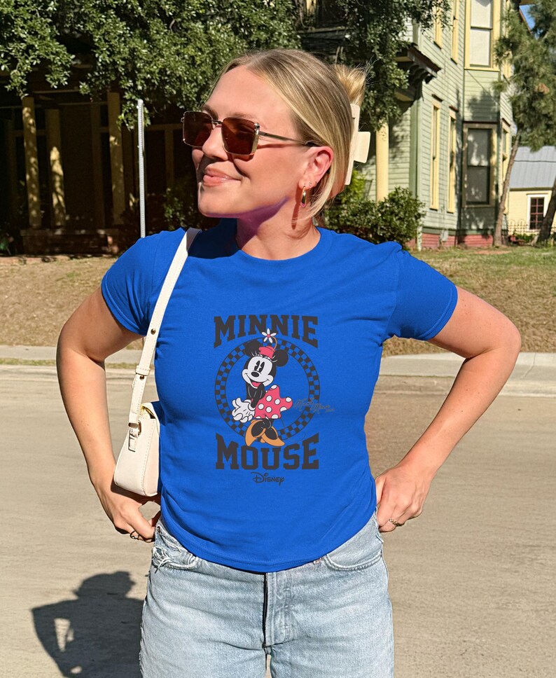 K&ouml;nnte beinhalten: Ein k&ouml;nigsblaues T-Shirt mit einem Minnie Mouse-Motiv. Das Design enth&auml;lt den Text "MINNIE MOUSE" in Schwarz, mit Minnie in einem gepunkteten Kleid. Das T-Shirt ist kurz&auml;rmelig und wird von einer Person getragen.