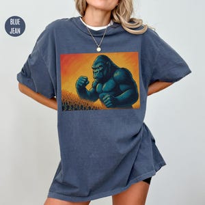May include: A blue jean oversized t-shirt with a graphic print of a large gorilla standing in front of a crowd of people.