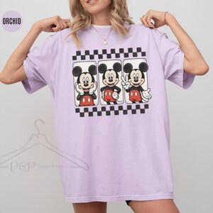 Puede incluir: Camiseta lila claro con tres ilustraciones de Mickey Mouse. Cada Mickey est&aacute; en una pose diferente, con pantalones cortos rojos y guantes blancos. El dise&ntilde;o est&aacute; enmarcado por un patr&oacute;n de tablero de ajedrez en blanco y negro.