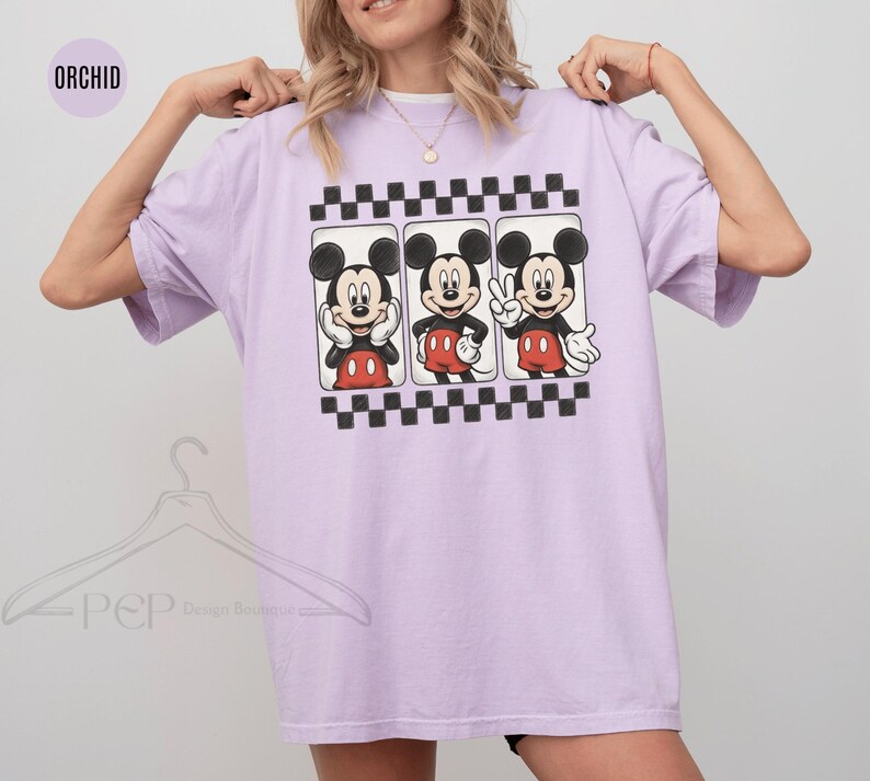Puede incluir: Camiseta lila claro con tres ilustraciones de Mickey Mouse. Cada Mickey est&aacute; en una pose diferente, con pantalones cortos rojos y guantes blancos. El dise&ntilde;o est&aacute; enmarcado por un patr&oacute;n de tablero de ajedrez en blanco y negro.