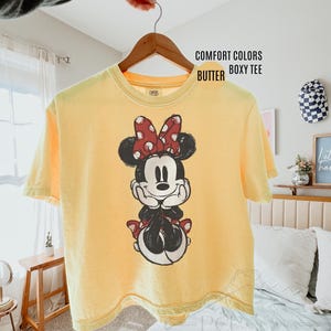 May include: A yellow Comfort Colors boxy tee featuring a Minnie Mouse graphic. The graphic shows Minnie Mouse in black, white, and red. The shirt is hanging on a wooden hanger.