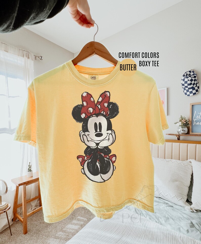 May include: A yellow Comfort Colors boxy tee featuring a Minnie Mouse graphic. The graphic shows Minnie Mouse in black, white, and red. The shirt is hanging on a wooden hanger.