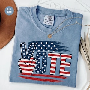 Op de afbeelding: Een blauw spijkerkleurig t-shirt met een afbeelding van een hand die een vredesteken maakt en het woord VOTE. Het ontwerp bevat het patroon van de Amerikaanse vlag met rode en witte strepen en witte sterren op een blauwe achtergrond.