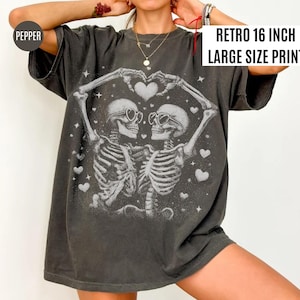 May include: Dark gray t-shirt with a large print featuring two skeletons embracing, forming a heart shape with their arms. The skeletons wear sunglasses and are surrounded by hearts and stars. The text "RETRO 16 INCH LARGE SIZE PRINT" is displayed on the upper right.