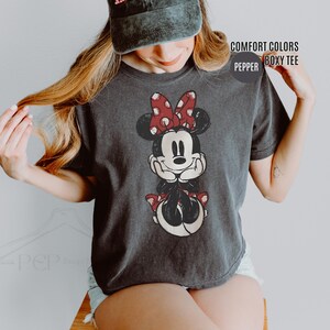 May include: A charcoal gray t-shirt featuring a distressed Minnie Mouse graphic. Minnie Mouse is depicted with a red polka dot bow, black ears, and a red polka dot skirt. The shirt has the words "COMFORT COLORS" and "BOXY TEE" printed on it.