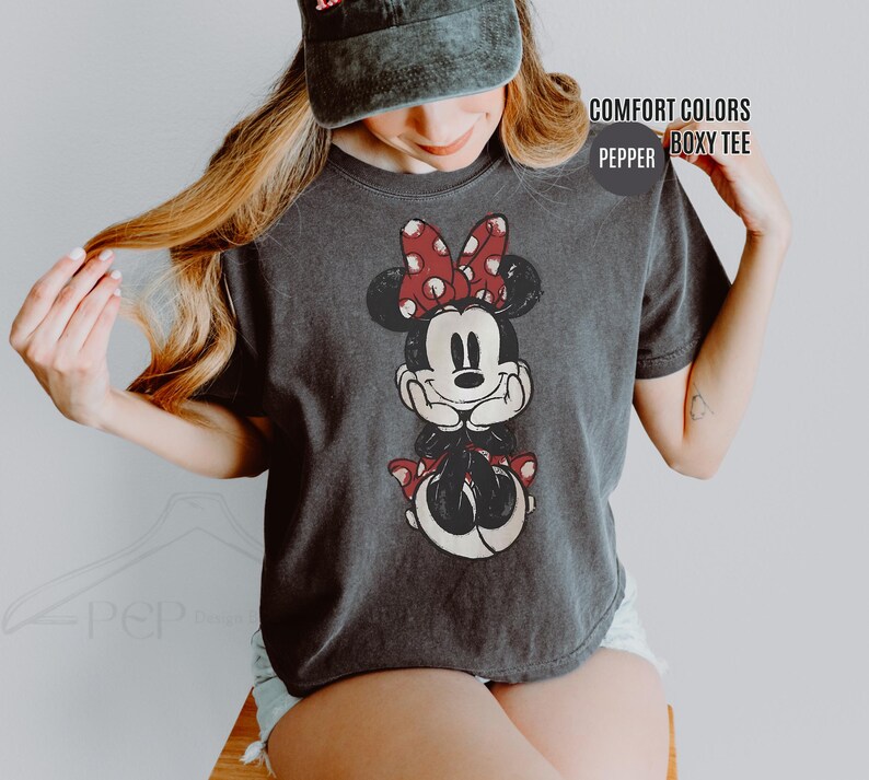 May include: A charcoal gray t-shirt featuring a distressed Minnie Mouse graphic. Minnie Mouse is depicted with a red polka dot bow, black ears, and a red polka dot skirt. The shirt has the words "COMFORT COLORS" and "BOXY TEE" printed on it.