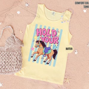 K&ouml;nnte beinhalten: Hellgelbes Tanktop mit dem Schriftzug "HOLD YOUR" in Pink und Blau und einer Cartoon-Pferdegrafik. Das Tanktop ist mit "Butter" und "Comfort Colors Tank Top" beschriftet. Links ist eine beige geh&auml;kelte Tasche.