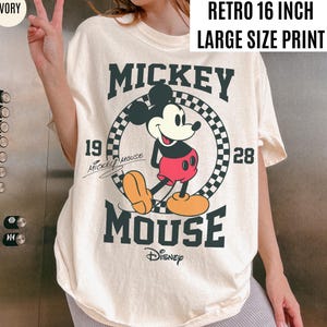 May include: Ivory-colored t-shirt featuring a large graphic of Mickey Mouse. The design includes the text "Mickey Mouse" in bold letters, the year "1928", and the Disney logo. The shirt has a "Retro 16 inch Large Size Print" label.