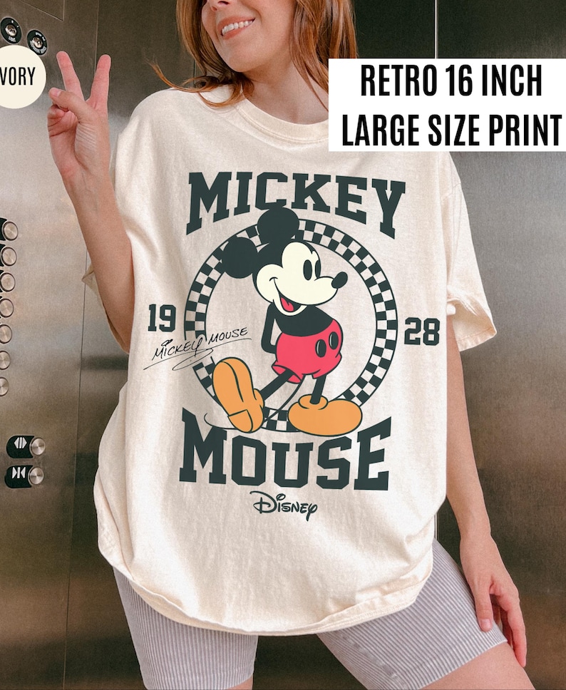May include: Ivory-colored t-shirt featuring a large graphic of Mickey Mouse. The design includes the text "Mickey Mouse" in bold letters, the year "1928", and the Disney logo. The shirt has a "Retro 16 inch Large Size Print" label.