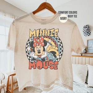 Może przedstawiać: Kremowa koszulka typu boxy z grafiką Myszki Minnie. Projekt przedstawia Minnie z czerwoną kokardą na tle w szachownicę. Napis "MINNIE MOUSE" jest w stylu retro. Koszulka wisi na drewnianym wieszaku.