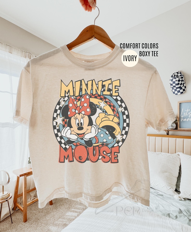 Może przedstawiać: Kremowa koszulka typu boxy z grafiką Myszki Minnie. Projekt przedstawia Minnie z czerwoną kokardą na tle w szachownicę. Napis "MINNIE MOUSE" jest w stylu retro. Koszulka wisi na drewnianym wieszaku.