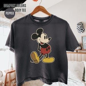May include: A dark grey boxy tee shirt with a vintage Mickey Mouse design. The cartoon mouse is wearing red shorts, yellow shoes, and white gloves. The shirt has the text "COMFORT COLORS BOXY TEE" and "PEPPER".