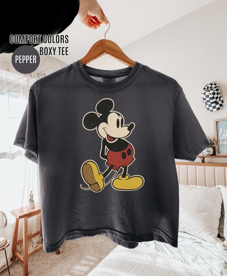 May include: A dark grey boxy tee shirt with a vintage Mickey Mouse design. The cartoon mouse is wearing red shorts, yellow shoes, and white gloves. The shirt has the text "COMFORT COLORS BOXY TEE" and "PEPPER".