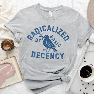 May include: A light grey t-shirt featuring the words "RADICALIZED BY BASIC DECENCY" in blue, and a blue bird graphic. The shirt is knotted at the bottom.