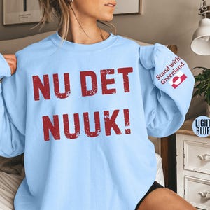 May include: Light blue sweatshirt with the words "NU DET NUUK!" in distressed red lettering across the front. The right sleeve features the text "Stand with Greenland" and a Greenlandic flag. The sweatshirt is a light blue color.
