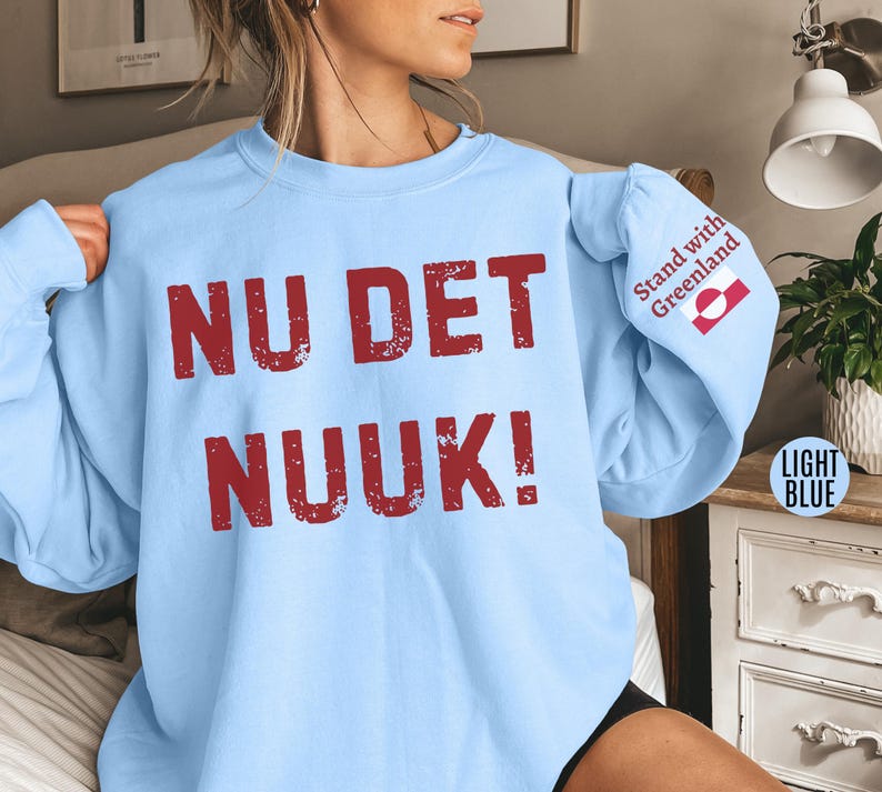 May include: Light blue sweatshirt with the words "NU DET NUUK!" in distressed red lettering across the front. The right sleeve features the text "Stand with Greenland" and a Greenlandic flag. The sweatshirt is a light blue color.