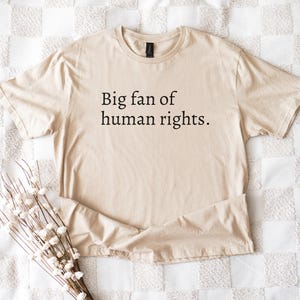 May include: A beige t-shirt with the text "Big fan of human rights" in black. The shirt is folded on a white and beige patterned surface, with a bundle of white flowers in the lower left corner.