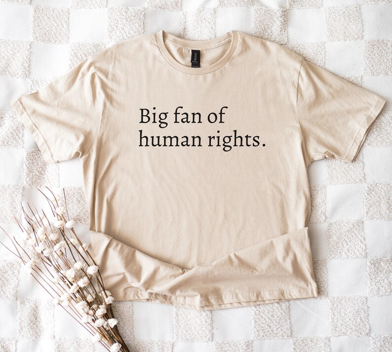 May include: A beige t-shirt with the text "Big fan of human rights" in black. The shirt is folded on a white and beige patterned surface, with a bundle of white flowers in the lower left corner.